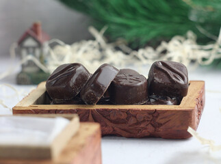 Chocolate candies lie in a wooden box on the table. Behind a spruce branch and a souvenir house.