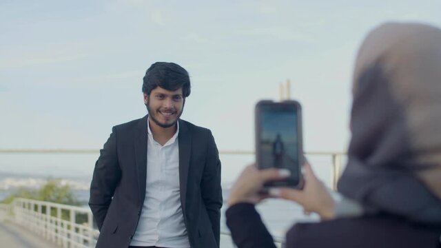 Girlfriend taking picture of her boyfriend on phone in city. Medium shot of beautiful Arabic couple having outdoor date. Handsome bearded man in official suit posing for camera. Love concept