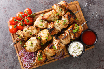 Delicious chicken kebabs on skewers with fresh vegetables and two sauces close-up on a wooden tray on a gray concrete background. horizontal top view from above