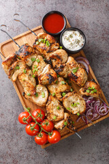 Homemade chicken kebab on skewers with vegetables and two sauces close-up in a wooden board on gray concrete background. Vertical top view from above