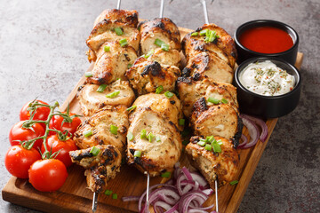 Juicy grilled chicken skewers with fresh vegetables and two sauces close-up on a wooden tray on a gray concrete background. horizontal