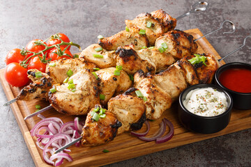 Delicious chicken kebabs on skewers with fresh vegetables and two sauces close-up on a wooden tray on a gray concrete background. horizontal