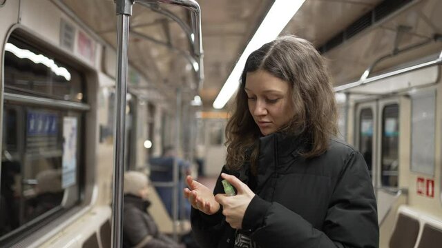 Young squeamish woman with dark wavy hair wearing warm jacket uses sanitizer on hands after holding handrail in underground carriage