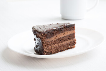 Chocolate cake is close-up on a white plate and blurred background with a white mug of coffee. Ideal dessert concept - 478371586