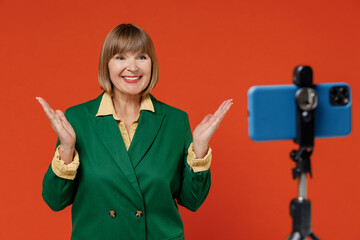 Elderly smiling happy woman 50s wear green classic suit talk by video call using mobile cell phone on stand spread hands isolated on plain orange color background. People business lifestyle concept.
