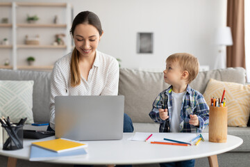 Adorable toddler boy drawing next to mom working on laptop, lady spending time with son and enjoying freelance work