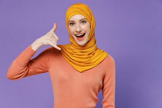 Smiling young arabian asian muslim woman in abaya hijab yellow clothes doing phone gesture like says call me back keep mauth wide open isolated on plain pastel light violet background studio portrait.
