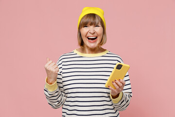 Elderly smiling happy fun woman 50s in striped shirt yellow hat hold using mobile cell phone do winner gesture isolated on plain pastel light pink background studio portrait. People lifestyle concept.