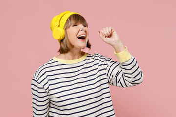 Elderly stylish woman 50s wearing striped shirt yellow hat headphones listen to music sing song in microphone isolated on plain pastel light pink background studio portrait. People lifestyle concept.