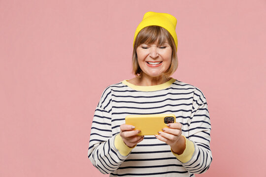 Elderly Fun Woman 50s In Striped Shirt Yellow Hat Using Play Racing App On Mobile Cell Phone Hold Gadget Smartphone For Pc Video Games Isolated On Plain Pastel Light Pink Background Studio Portrait