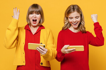 Winner woman 50s in red shirt with girl 12-13 years old. Grandmother granddaughter using play racing app on mobile cell phone hold gadget smartphone pc video games isolated on plain yellow background