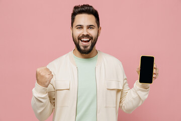 Young smiling happy man 20s wearing trendy jacket shirt hold in hand use mobile cell phone browsing with blank screen workspace area do winner gesture isolated on plain pastel light pink background.