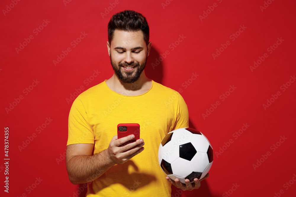 Wall mural Cheerful young bearded man football fan in yellow t-shirt cheer up support favorite team hold soccer ball use mobile cell phone read text chat isolated on plain dark red background studio portrait