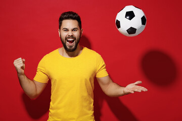 Exultant fun young bearded man football fan in yellow t-shirt cheer up support favorite team tossed soccer ball celebrate clenching fists say yes isolated on plain dark red background studio portrait.