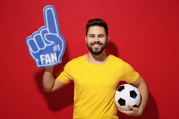 Blithesome young bearded man football fan in yellow t-shirt cheer up support favorite team look camera hold soccer ball fan foam glove finger up isolated on plain dark red background studio portrait. - obrazy, fototapety, plakaty