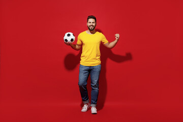 Fototapeta na wymiar Full size body length young bearded man football fan in yellow t-shirt cheer up support favorite team hold soccer ball demonstrate strength power isolated on plain dark red background studio portrait.