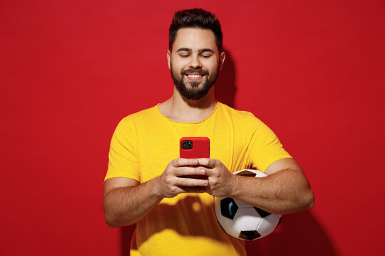 Cheerful happy smiling fun young bearded man football fan in yellow t-shirt cheer up support favorite team hold soccer ball use mobile cell phone isolated on plain dark red background studio portrait.