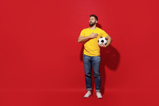 Full size body length happy young bearded man football fan in yellow t-shirt support favorite team hold soccer ball sing national country anthem isolated on plain dark red background studio portrait.