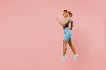 Full body young strong sporty fitness trainer instructor woman wear blue tracksuit headphones spend time in home gym jump high run isolated on pastel plain light pink background Workout sport concept
