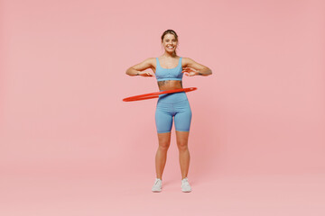 Full size young strong sporty athletic fitness trainer instructor woman wear blue tracksuit spend time in home gym using hula hoop isolated on pastel plain light pink background Workout sport concept.