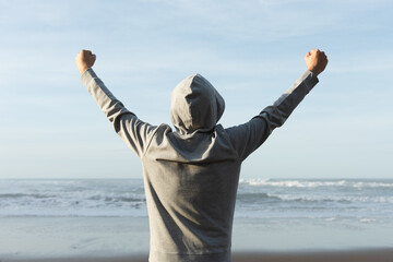 Rear view of man wearing a hoodie celebrating victory or good results while looking to the sea