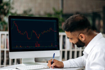 Smart successful concentrated confident Indian trader expert, stock broker, using computer in his modern office, analyzing cryptocurrency market, taking notes, planning stock strategy