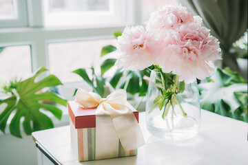 Fresh bouquet of tender pink peonies flowers and gift box with satin bow on white table with a light biophilic interior background. Festive flowers as gift. Mockup for greeting card design.