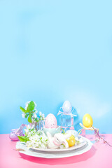 Easter table setting decoration, with colorful Easter eggs, spring garden blossom flower, bunny rabbits decor, on high-colored blue background flatlay copy space