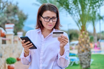 Middle age smiling woman with credit card and smart phone in hands, outdoor