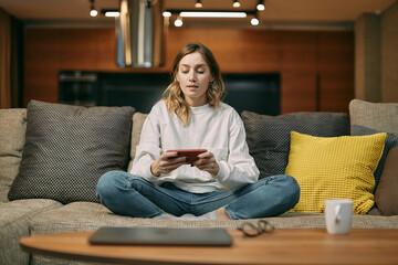 Female gamer playing mobile online game on smart phone with friends online sitting on sofa at home...