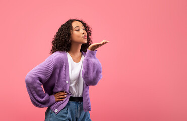 Lovely young black woman blowing air kiss at camera over pink studio background, copy space. Valentine's Day concept