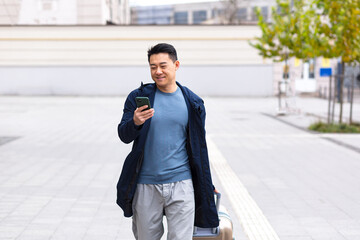 Asian tourist, male passenger arrives from the airport, walks around the city with a suitcase, a man smiles and rejoices uses the phone and the application to book accommodation and book a taxi