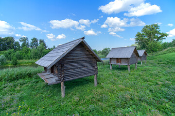 Three old Slavic wooden barns on poles near the river. Summer. Blue sky with clouds. Daylight.
