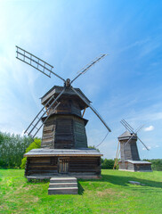 Two old wooden windmills made of logs on green grass. There is a forest in the background. Summer. Clear blue sky with clouds. Daylight.