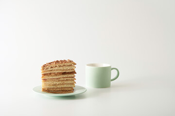 A piece of layered homemade honey cake with custard on a plate next to a cup on a white background. Copy space.