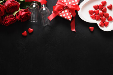 Valentine's day greeting card with gift, chocolate sweets, red roses, and red wine with on black background. View from above. Copy space.