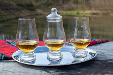 Tasting glasses of scotch whisky strong drink and red Scottisch tartan on old outdoor table with forest lake on background