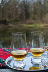 Tasting glasses of scotch whisky strong drink and red Scottisch tartan on old outdoor table with forest lake on background
