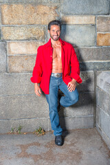 Obraz premium Dressing in a red long sleeve shirt, unbuttoned, blue jeans, leather shoes, a sexy, middle age guy with mustache and beard is leaning against the wall, smilingly looking at you...