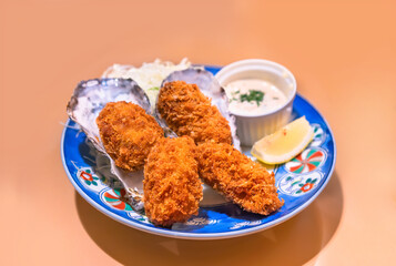 Japanese shellfish dish of battered and deep fried oysters made by adding egg whites and bread crumbs called kaki-furai or kaki fry served in their shell with a sauce and a lemon slice in a plate.