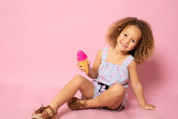 Obraz na płótnie Canvas Cute little girl in summer clothing with ice cream sitting on the floor isolated over pink background.
