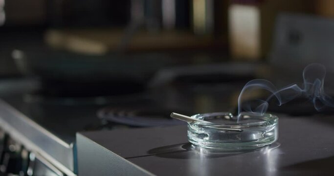 Interior detail shot of ashtray that has a lit marijuana CBD pre-roll joint, cigarette with smoke rising sitting in kitchen next to stove top