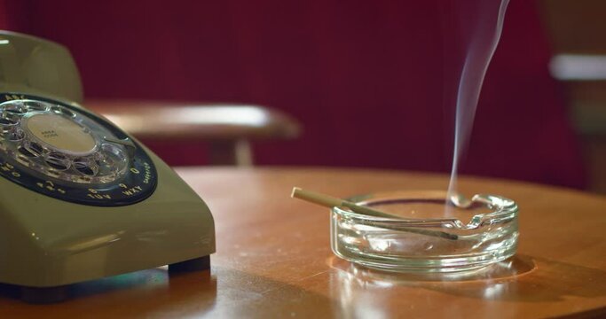 Home Interior detail shot of side table with analog vintage rotary telephone and ashtray that has a lit cannabis pre-roll joint, cigarette with smoke rising