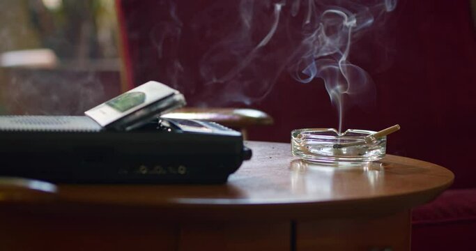 Low angle home interior detail shot of side table with vintage tape recorder / answering machine and ashtray that has a lit CBD marijuana pre-roll joint, cigarette with smoke rising
