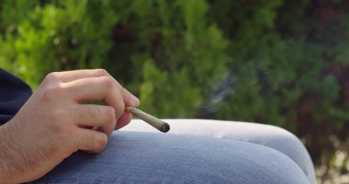 Eye level detail shot of man holding a lit marijuana CBD pre-roll cannabis joint, cigarette with smoke rising sitting outdoors