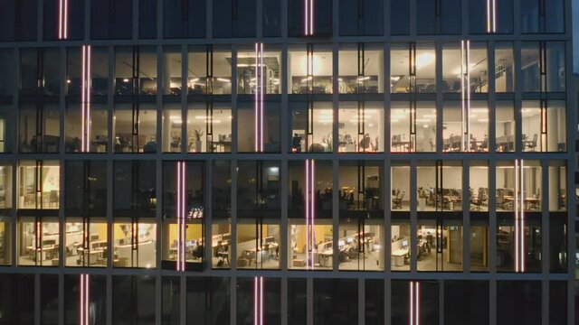 Cinematic drone shot at offices with people working on computers. Zooming in on the windows of a skyscraper. Office block at night revealing the daily activities of office workers working late