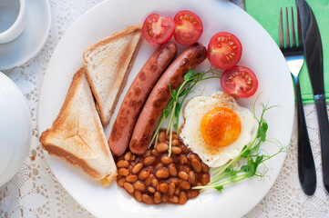 English breakfast with scrambled egg, sausages, toast, tomatoes, pea sprouts and beans on white plate. Traditional english breakfast with tea and teapot on white lace tablecloth. Top view