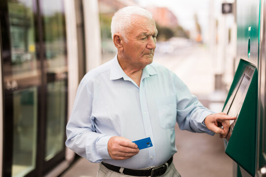 Senior man using ATM machine with credit card while waiting for tram on station.