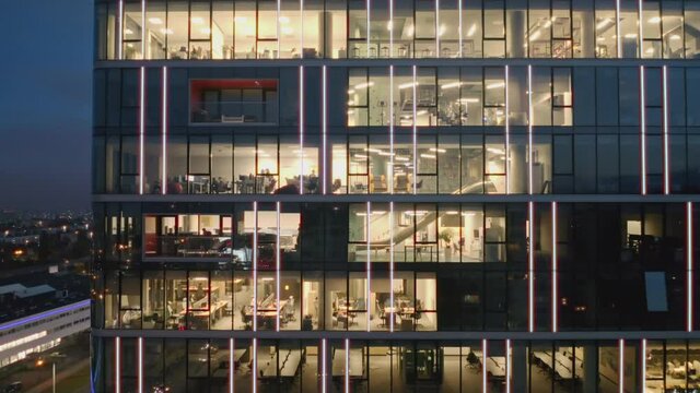 Aerial view of the night windows of offices in a modern skyscraper with business people working late. Movement of the camera panning from top to bottom on the windows of offices at night.