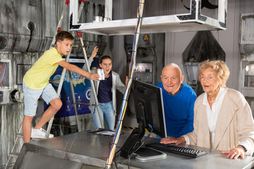 Fototapeta na wymiar Senior couple using computer while standing in escape room. Teenage girl shouting at them, young boy standing on stepladder with can in hand.